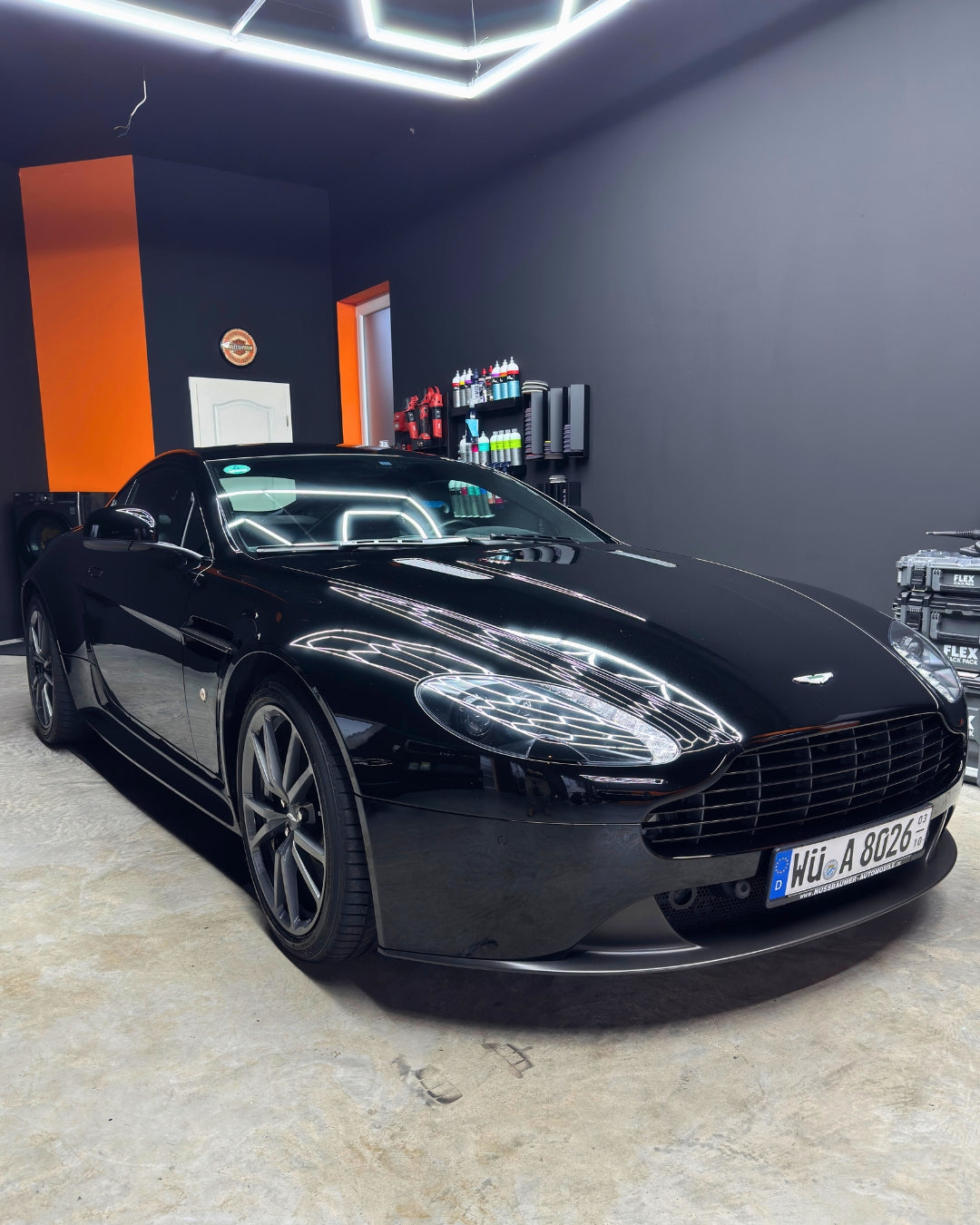Schwarzer Sportwagen von Aston Martin in einer modern beleuchteten Werkstatt mit orange-schwarzen Wänden.