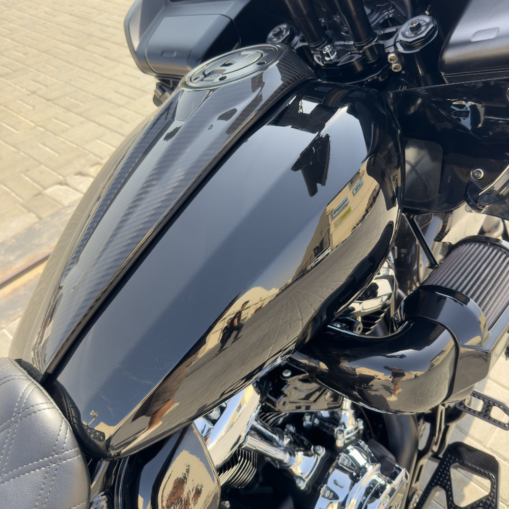 Glänzender schwarzer Motorradtank mit Carbonakzent und Spiegelung von Gebäuden und Himmel auf der Oberfläche.