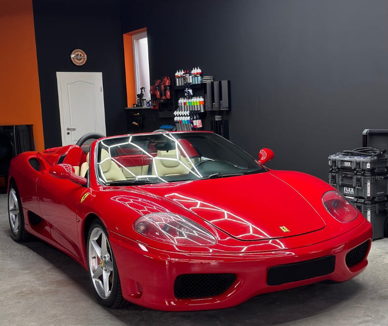 Roter Ferrari mit beigem Innenraum in einer modernen, sauber beleuchteten Werkstatt mit Pflegeprodukten und Werkzeugen im Hintergrund.“