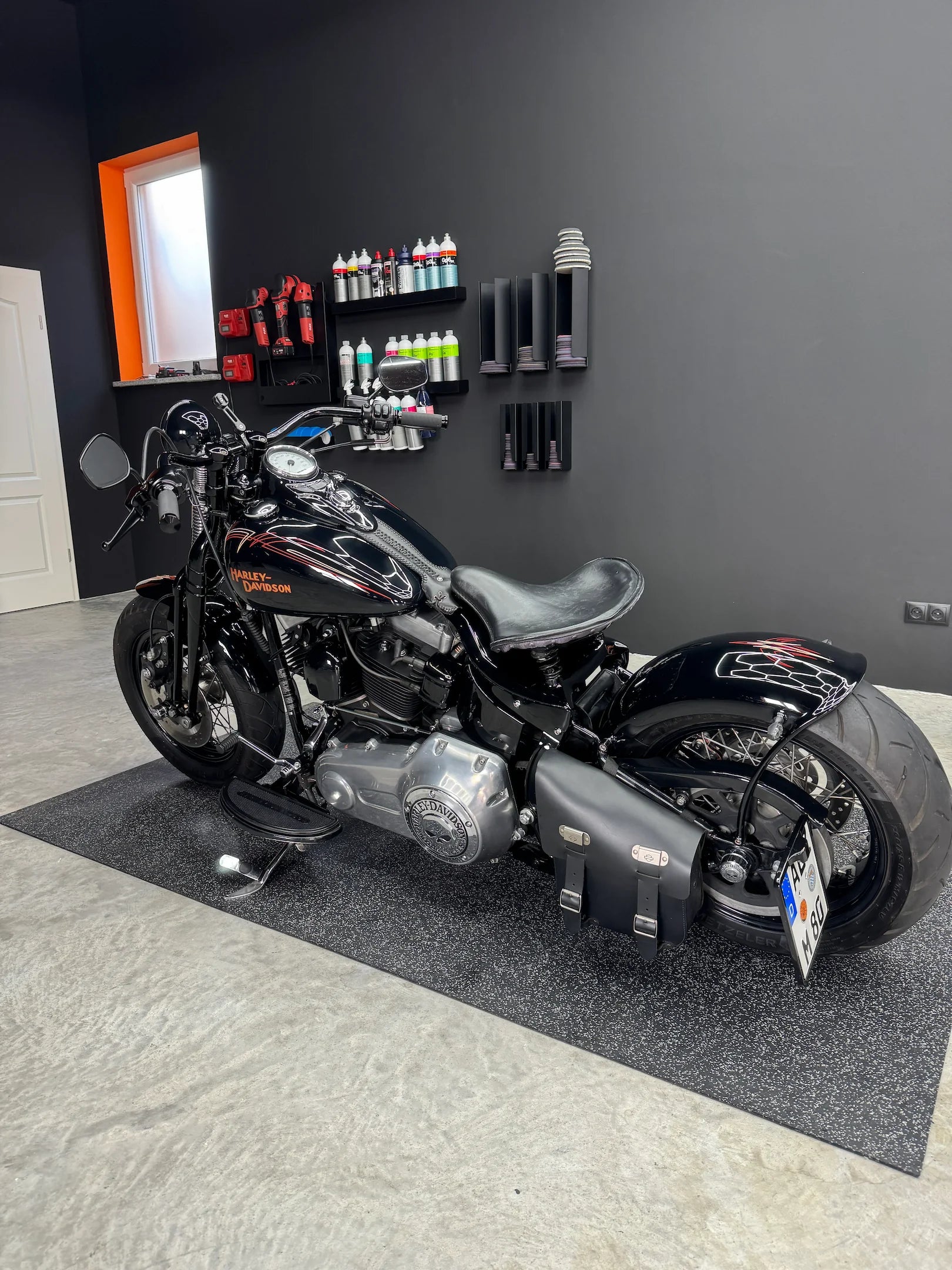 Schwarzes Harley-Davidson-Motorrad mit Ledertasche steht in einer gepflegten, modern eingerichteten Werkstatt mit Pflegeprodukten an der Wand.