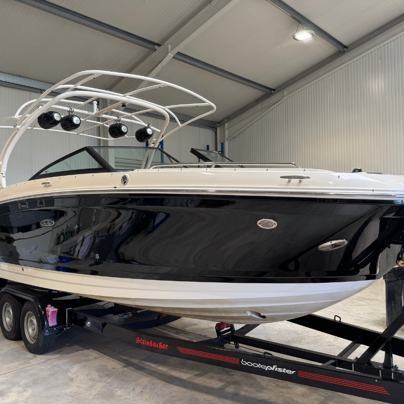 Schwarz-weißes Motorboot auf Anhänger in einer hellen Halle, mit glänzender Bordwand und montiertem Überrollbügel mit Lautsprechern.