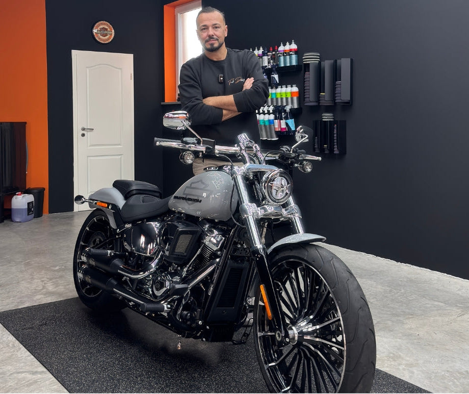 Ein Mann steht mit verschränkten Armen neben einem grauen Harley-Davidson-Motorrad in einer modernen Werkstatt mit Autopflege-Produkten an der Wand.